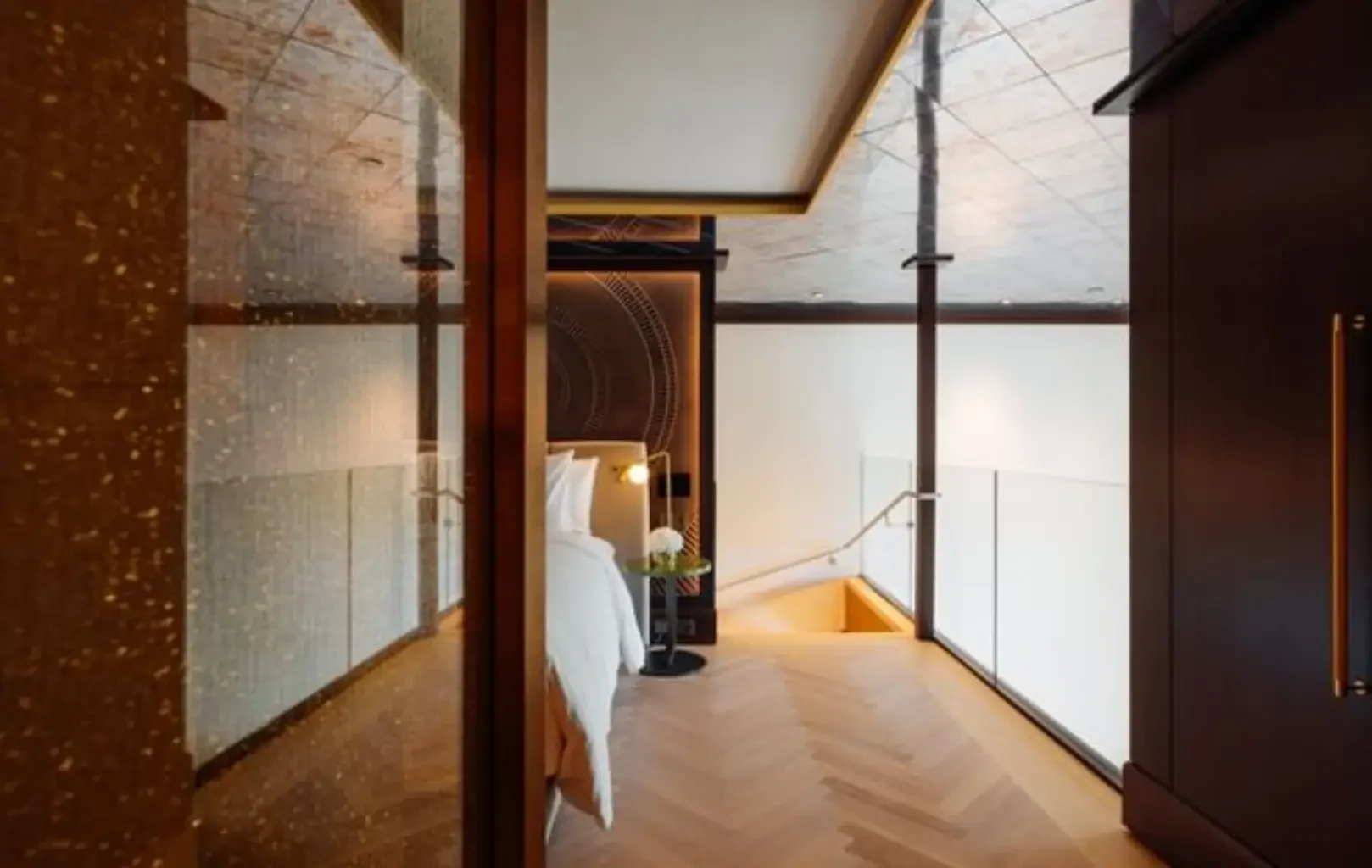 Ein modernes Loft-Schlafzimmer mit einem hübsch gemachten Bett, Holzfußboden, einem Glasgeländer an der Treppe und warmer Beleuchtung - perfekt für alle, die gerne vorausplanen und Wert auf durchdachtes Design legen.