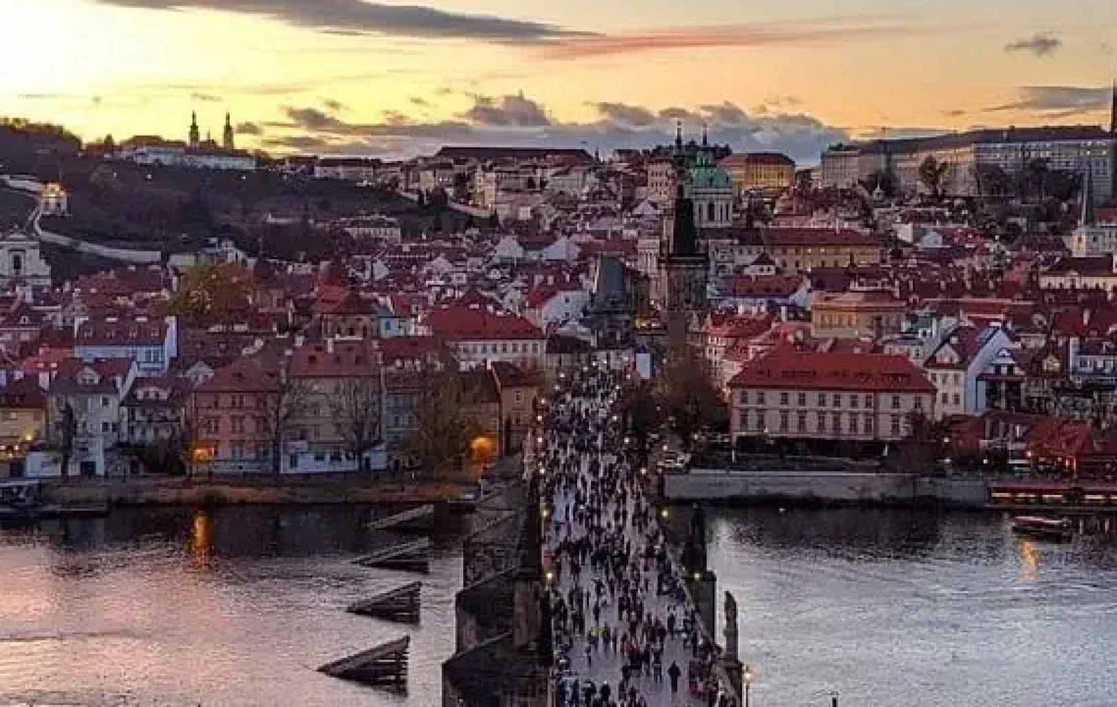 Blick auf eine überfüllte historische Steinbrücke über einen Fluss, mit rotgedeckten Gebäuden und Türmen im Hintergrund bei Sonnenuntergang.