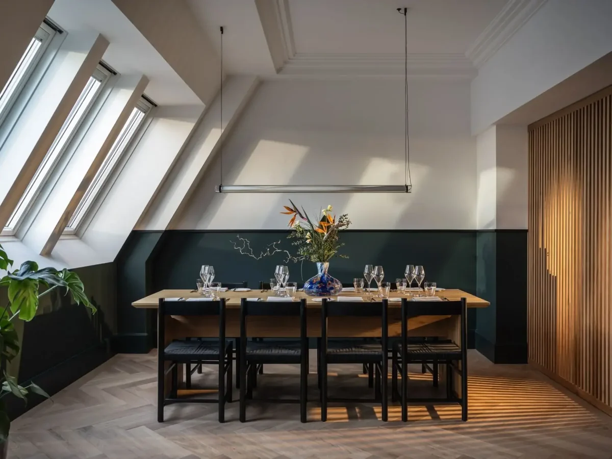 A modern dining room in the Presidential Suite features a wooden table set for eight, black chairs, a vase with flowers, angled skylights, and minimalist décor.