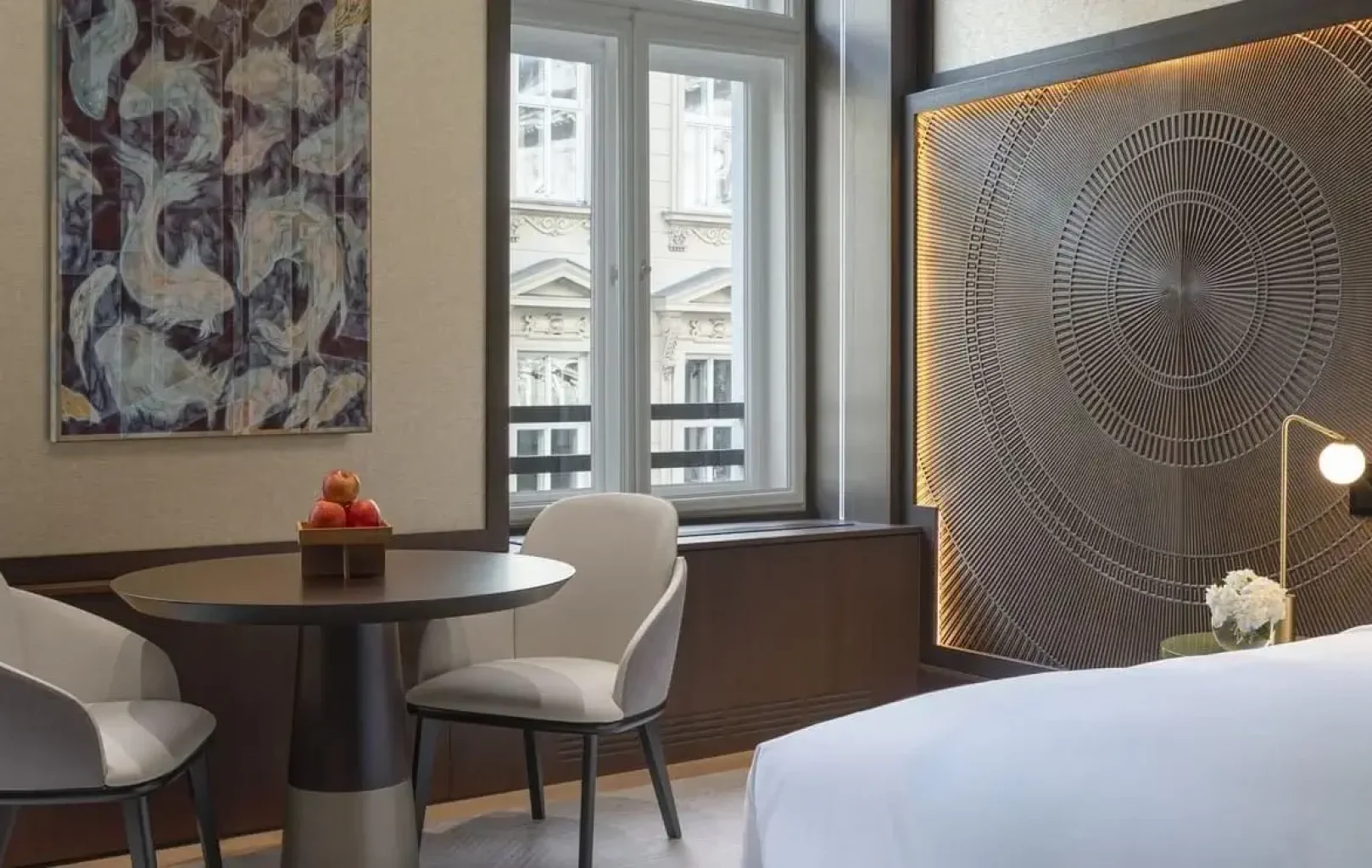 Modern hotel room with Summer Elegance, featuring a round table and two chairs by large windows, abstract wall art, and a bed with white linens and a geometric headboard design.