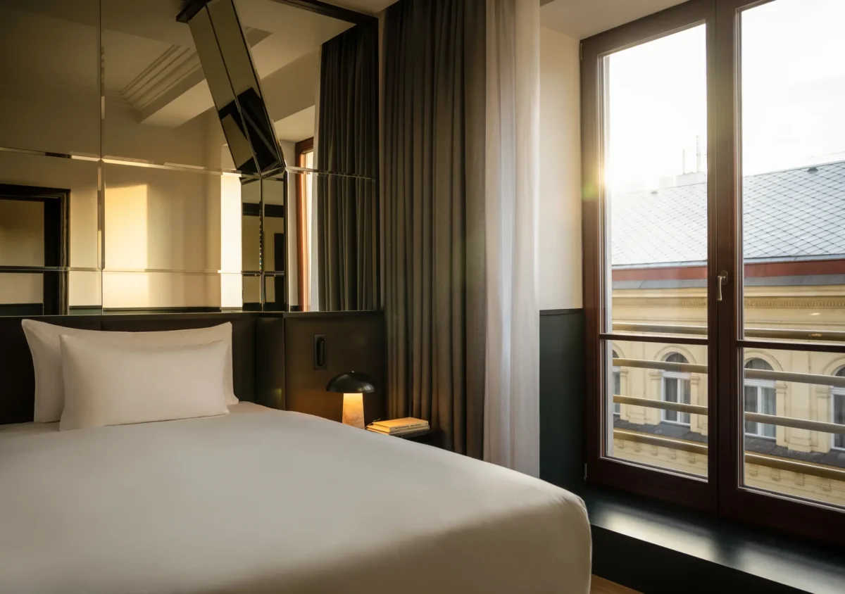 A modern Executive Suite featuring a neatly made bed, bedside lamp, mirrored wall panels, and large windows overlooking a building at sunset.