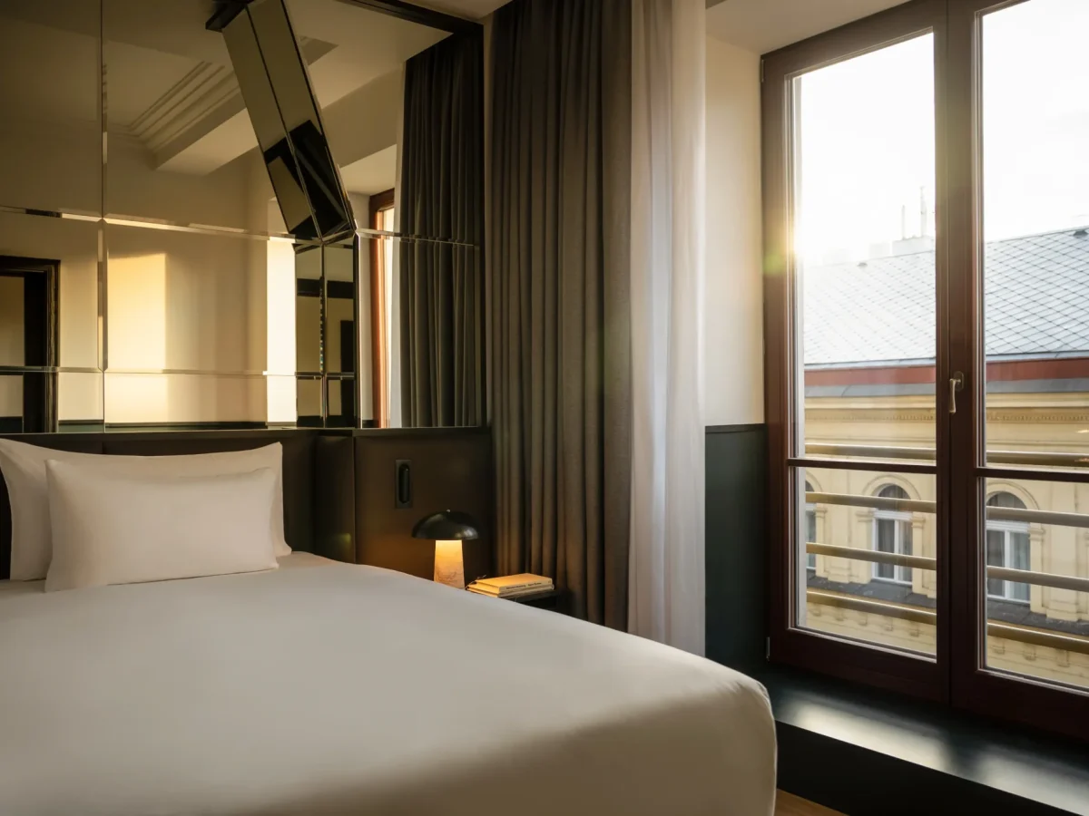 Moderní pokoj hotelu Apartmá Executive s úhledně ustlanou postelí, zrcadlovou stěnou, noční lampičkou a velkým oknem s výhledem na městskou zástavbu při západu slunce.
