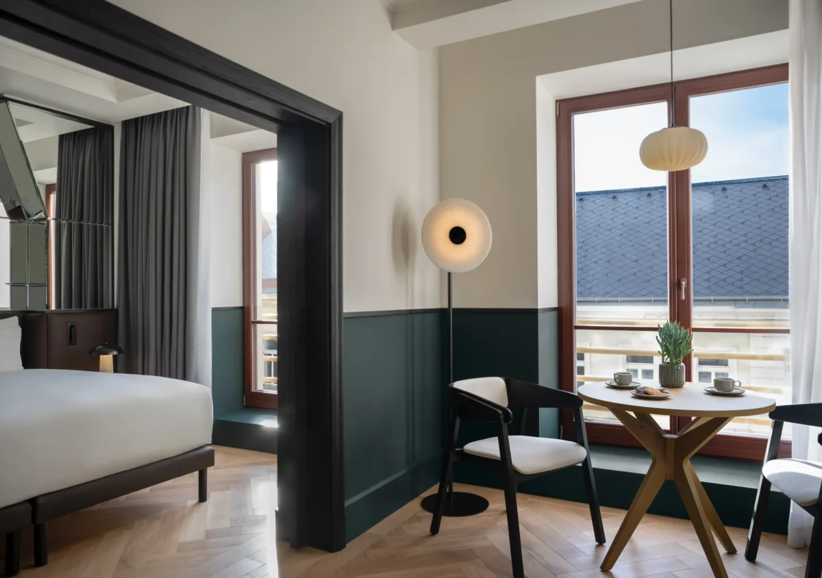 Modern Executive Suite featuring a bed, round table set for two, two chairs, large windows, and contemporary lighting. Natural light fills the space.