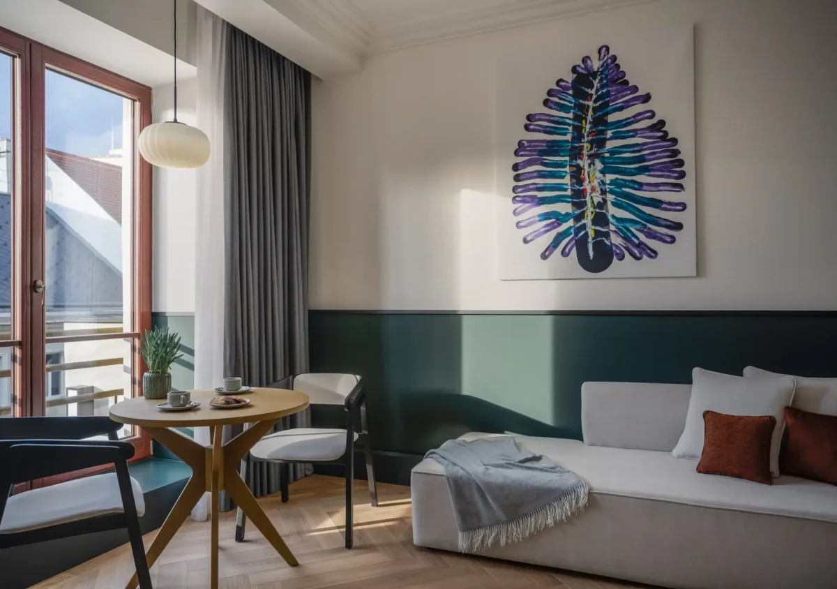 A modern executive suite living room with a light sofa, a small round dining table, two chairs, a large window, and abstract artwork on the wall above the sofa.