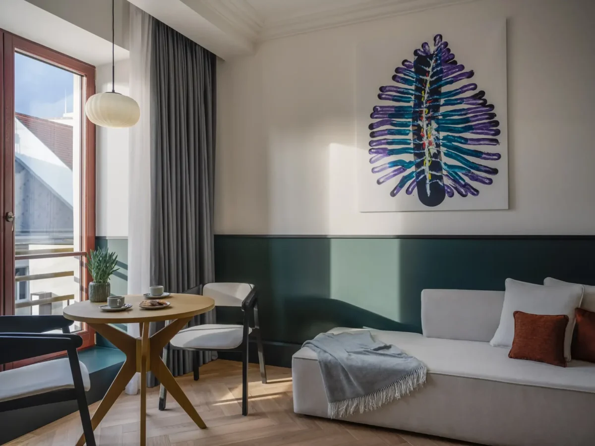 A modern executive suite living room with a light sofa, a small round dining table, two chairs, a large window, and abstract artwork on the wall above the sofa.