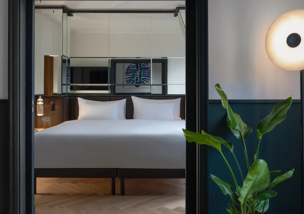 A modern Executive Suite featuring a double bed with white bedding, mirrored wall, indoor plant, round floor lamp, and elegant wood herringbone floor.