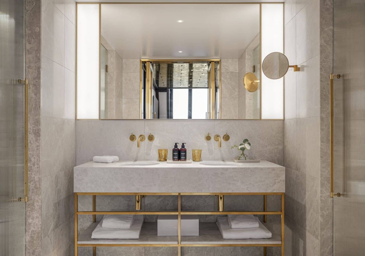 Modern bathroom with Almanac Deluxe double sink vanity, marble countertop, gold fixtures, large mirror, toiletries, towels on shelves, and neutral-toned tile walls.