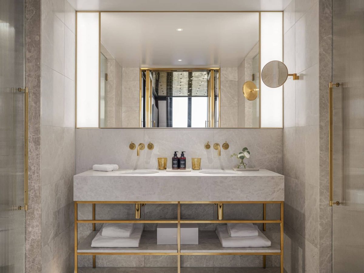 Modernes Badezimmer mit Doppelwaschbecken, Marmorarbeitsplatte, goldenen Armaturen, großem Spiegel und offenen Regalen für Handtücher und Toilettenartikel.