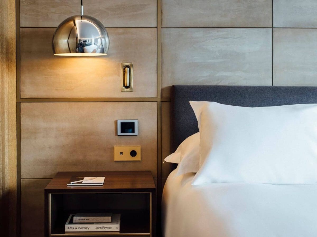 Una habitació d'hotel moderna de l'Habitació Gallery compta amb un llit ben fet, una tauleta de nit de fusta amb llibres i un bloc de notes, un llum de paret i un termòstat en una paret amb panells beix.