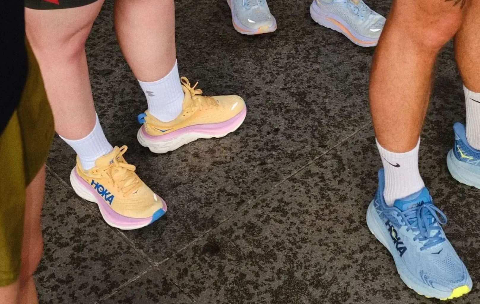Ein Laufclub steht auf dem strukturierten grauen Pflaster, ihre Hoka-Schuhe sind stilvoll aufgereiht - sie lieben sie für jeden Schritt. Nur Beine und Füße sind zu sehen, vereint durch Leidenschaft.