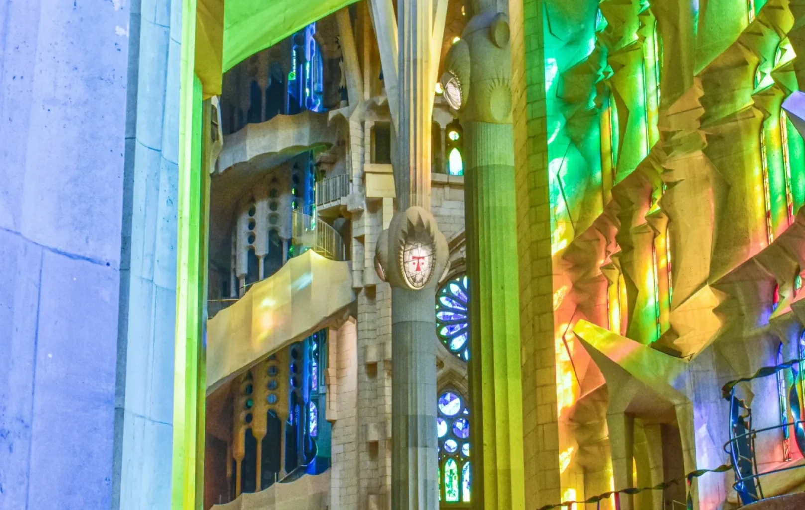 Vista interior de la Sagrada Familia con vidrieras de colores que proyectan luz verde y amarilla, altas columnas y visitantes de pie junto a la pared, un ambiente que recuerda al de las grandes salas de conciertos como L'Auditori de Barcelona.