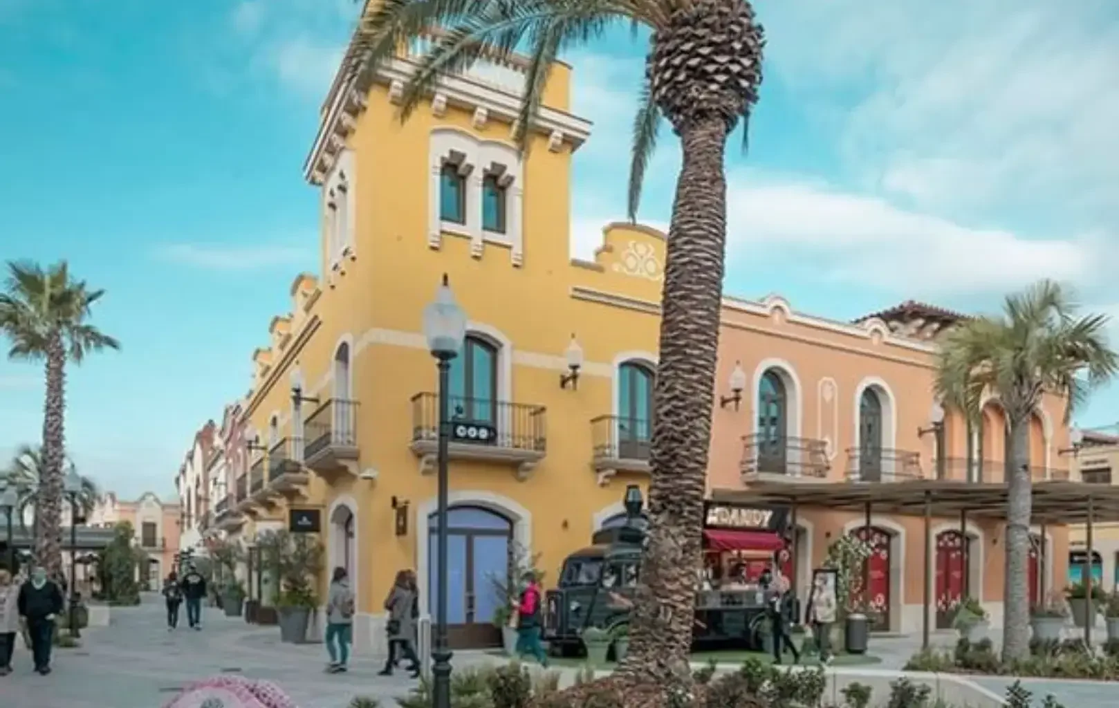 Una escena callejera con un edificio amarillo de estilo mediterráneo, palmeras, peatones disfrutando de una Escapada Shopping y tiendas al aire libre bajo un cielo parcialmente nublado.