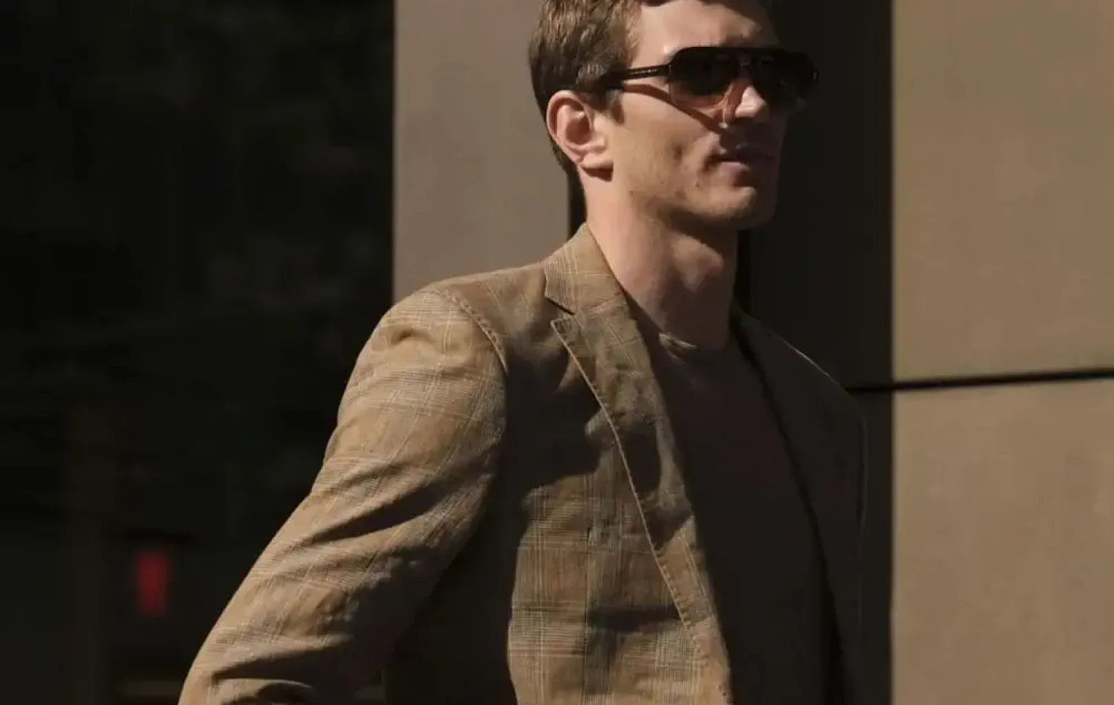 Un hombre con gafas de sol y un traje de cuadros escoceses se encuentra al aire libre con las manos en los bolsillos, parcialmente iluminado por la luz del sol, listo para una escapada de compras.