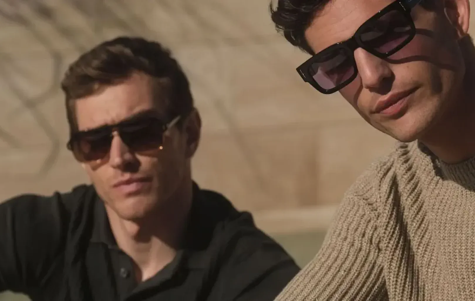 Dos hombres con gafas de sol se sientan al aire libre después de una Escapada Shopping; uno lleva un polo negro y el otro un jersey de punto beige.