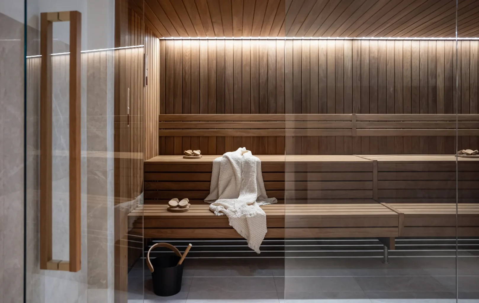 A modern wooden sauna room with benches, a towel and slippers on the seat, a bucket with a ladle on the floor, and soft overhead lighting—perfect for wellness and spa relaxation.