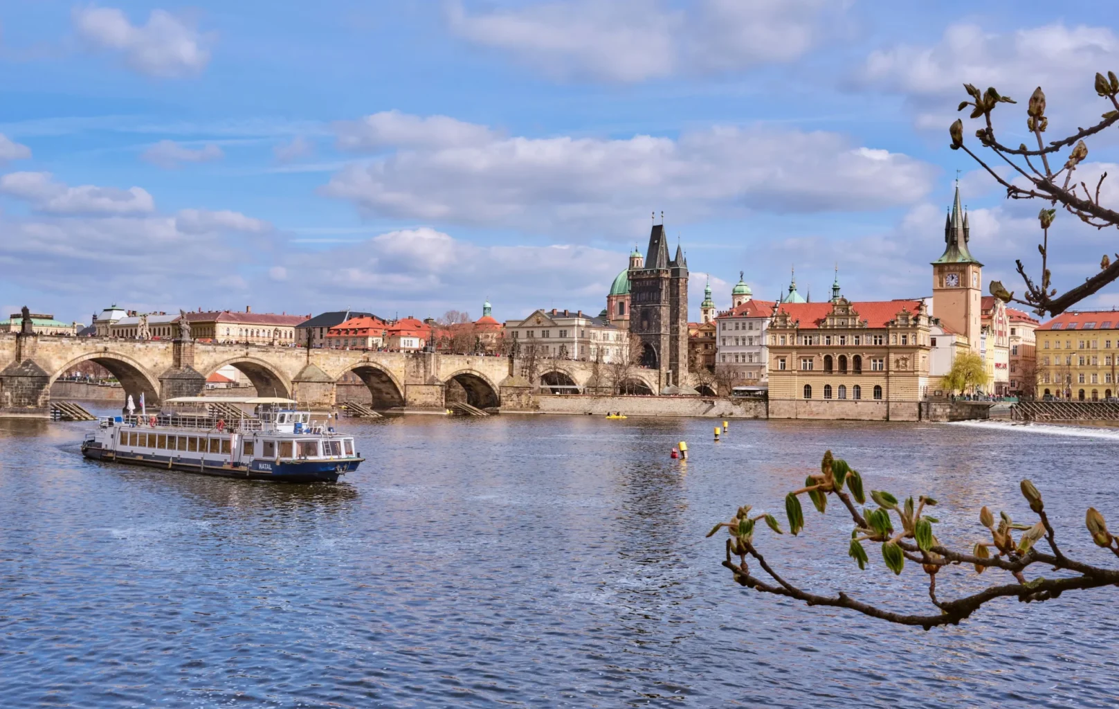 A sightseeing boat travels on a river near the historic Charles Bridge in Prague, with old buildings and towers in the background under a partly cloudy sky, just a short distance from bustling Wenceslas Square.