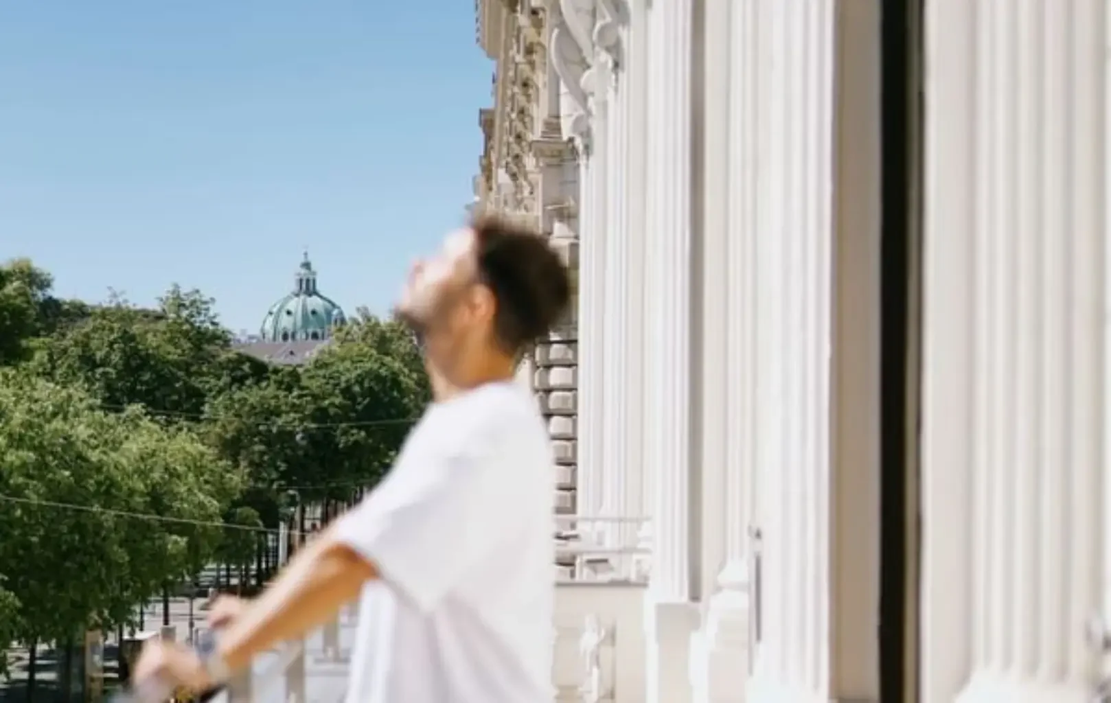 A person in a white shirt stands on a balcony, enjoying the Suite Experience, with blurred architectural columns and greenery in the background under a clear blue sky.