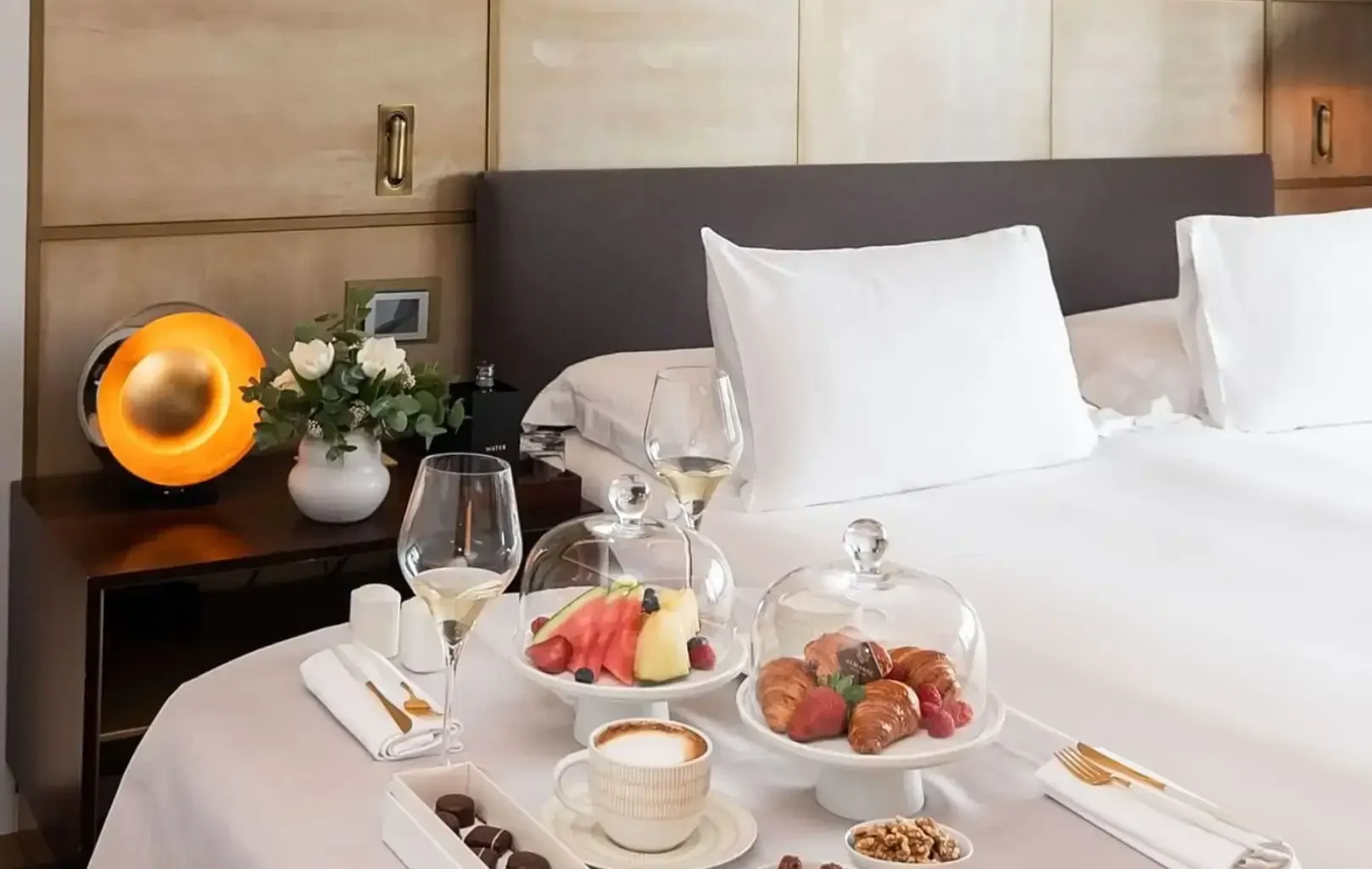 Una bandeja de desayuno con café, bollería, fruta, frutos secos y dos copas de vino está colocada sobre una cama pulcramente hecha, capturando la esencia de una refinada Experiencia Suite en una moderna habitación de hotel.