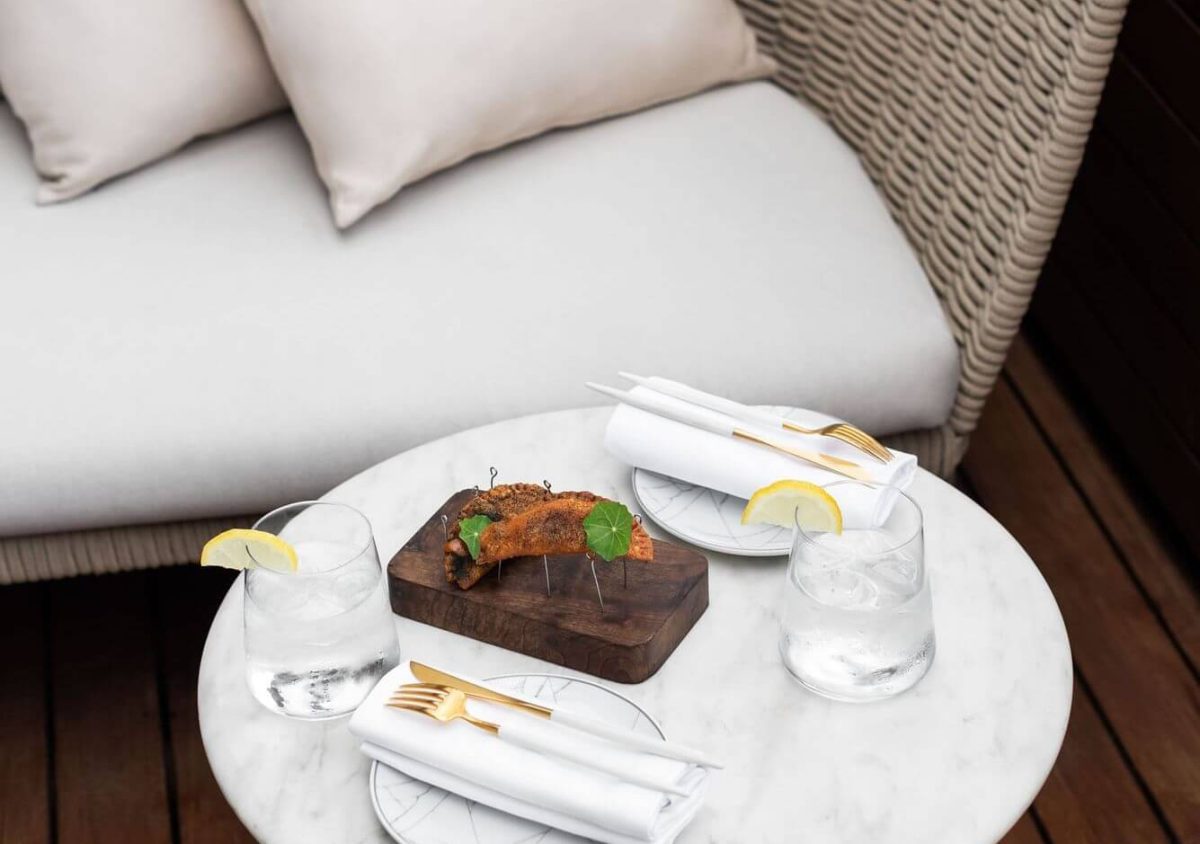 Una mesa de mármol con dos vasos de agua con limón, dos cubiertos y una tabla de madera con comida se encuentra junto a un sofá de mimbre acolchado en la elegante Suite Penthouse.