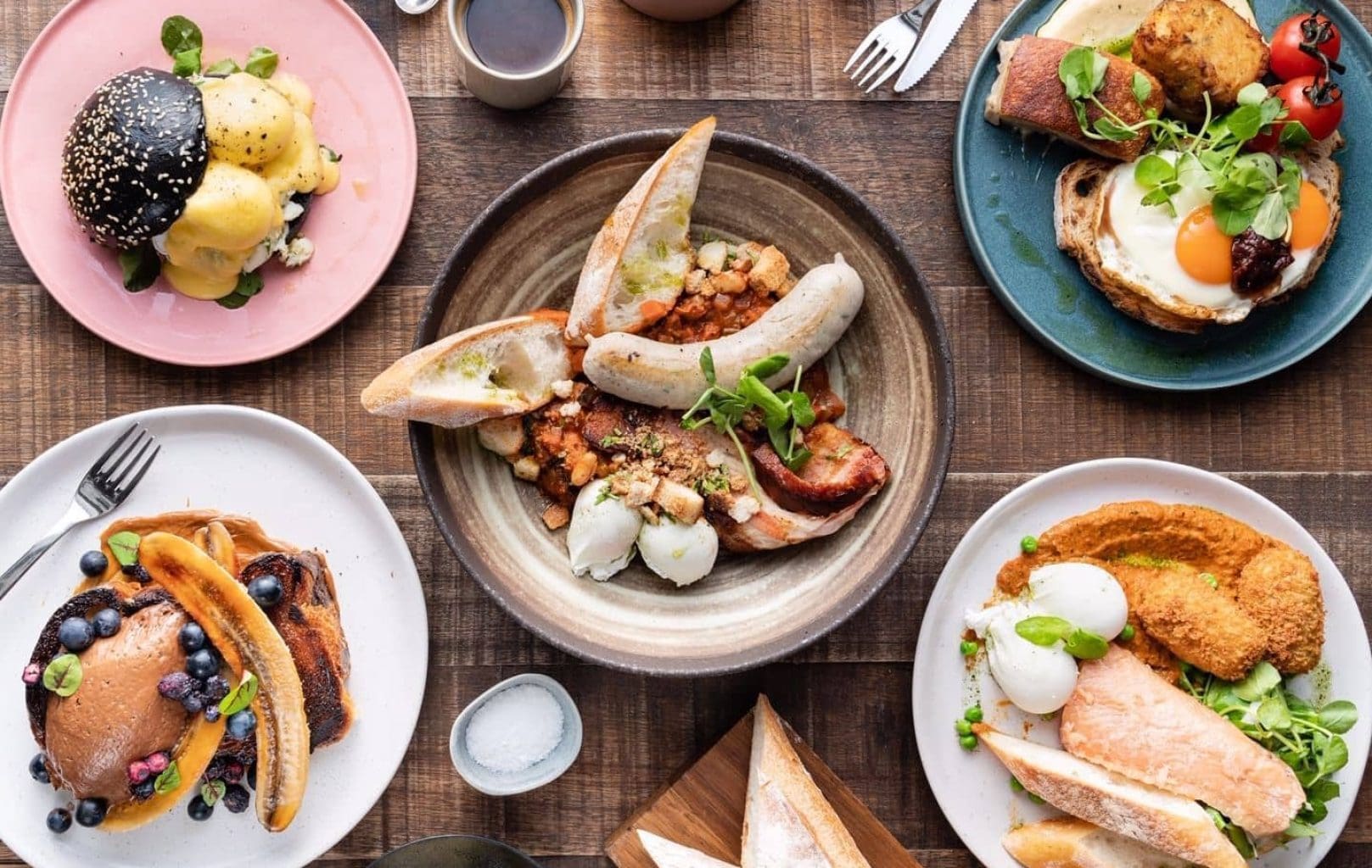 Una varietat de plats d'esmorzar amb ous, torrades, creps, salsitxes, verdures, salmó i cafè estan disposats sobre una taula de fusta a Hardware Société Barcelona.