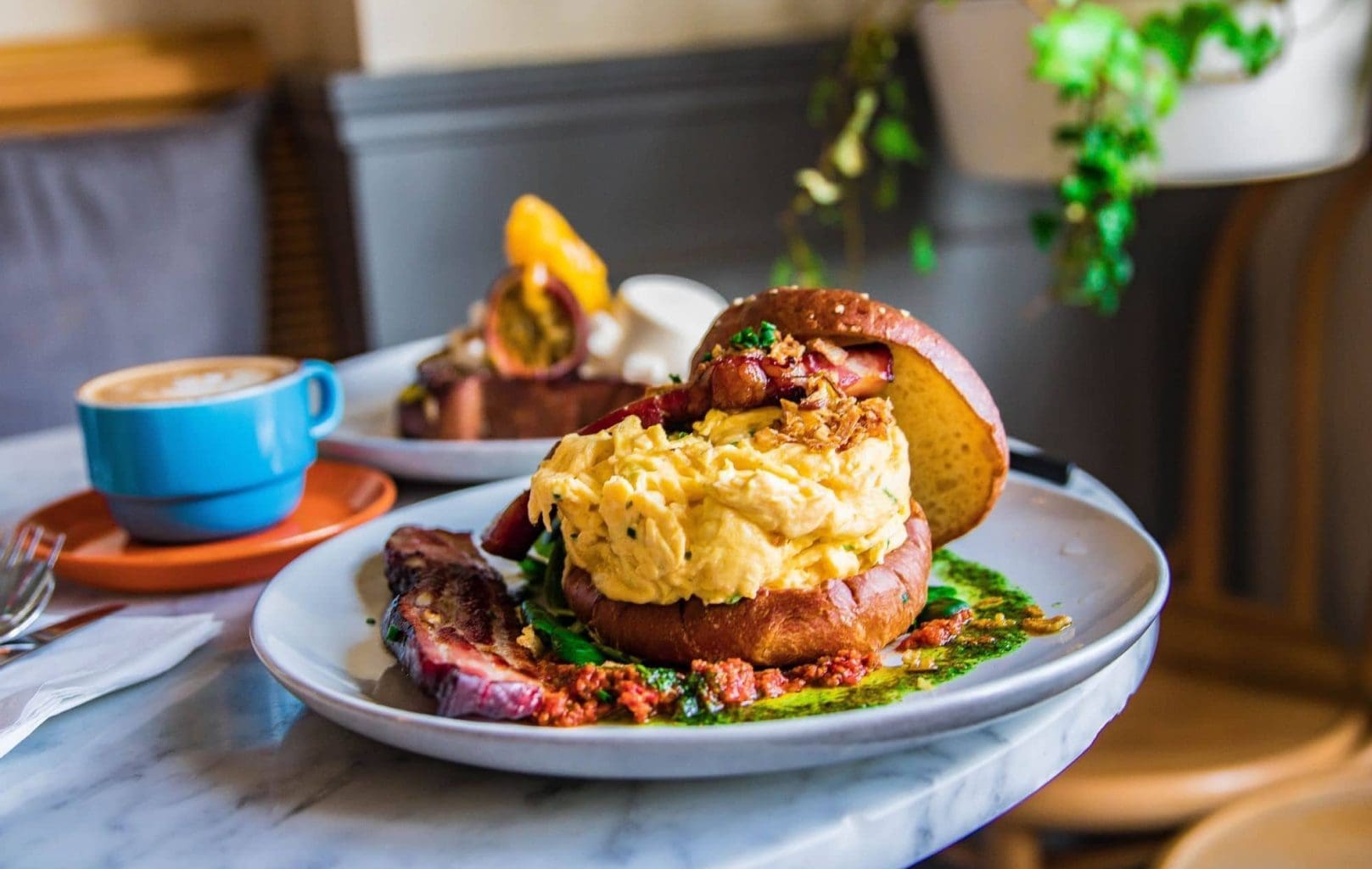 Un plat amb un entrepà d'ou remenat i bacó en un panet, servit amb salsa i verdures, es troba sobre una taula de marbre a Hardware Société Barcelona al costat d'una tassa de cafè blava.