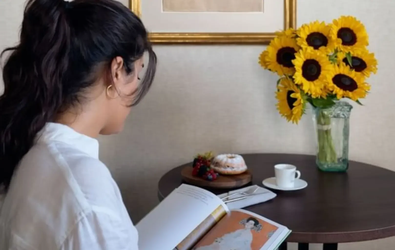 Eine Person in einem weißen Hemd sitzt an einem Tisch und liest in aller Ruhe ein Buch. Auf dem Tisch stehen eine Vase mit Sonnenblumen, eine Kaffeetasse, ein Teller mit Obst und ein Gebäck.