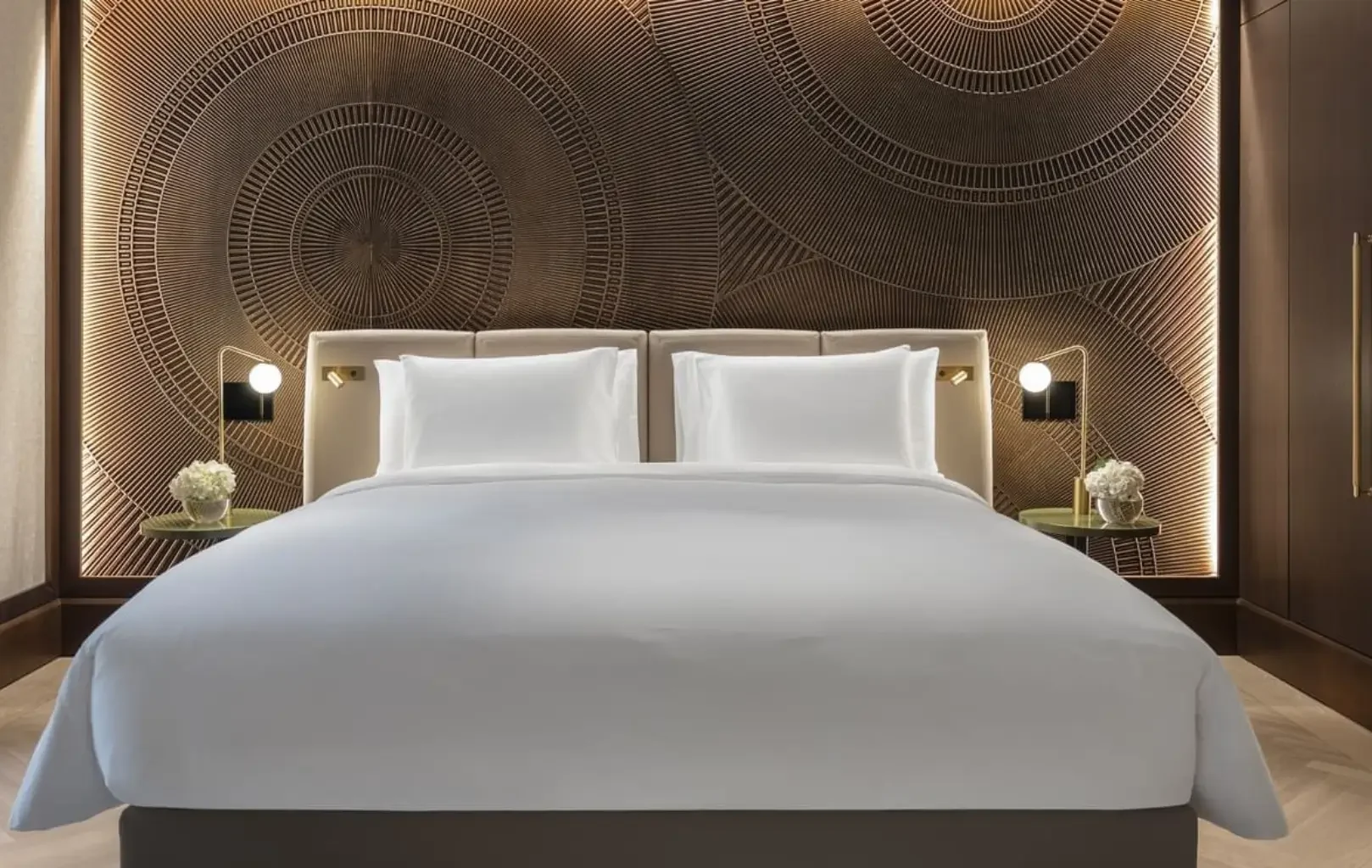 Ein modernes Hotelzimmer mit einem sauber gemachten Kingsize-Bett, zwei Nachttischen mit Lampen und Blumen und einer dekorativen, von Klimts Kunst inspirierten geometrischen Wandtafel hinter dem Kopfteil.