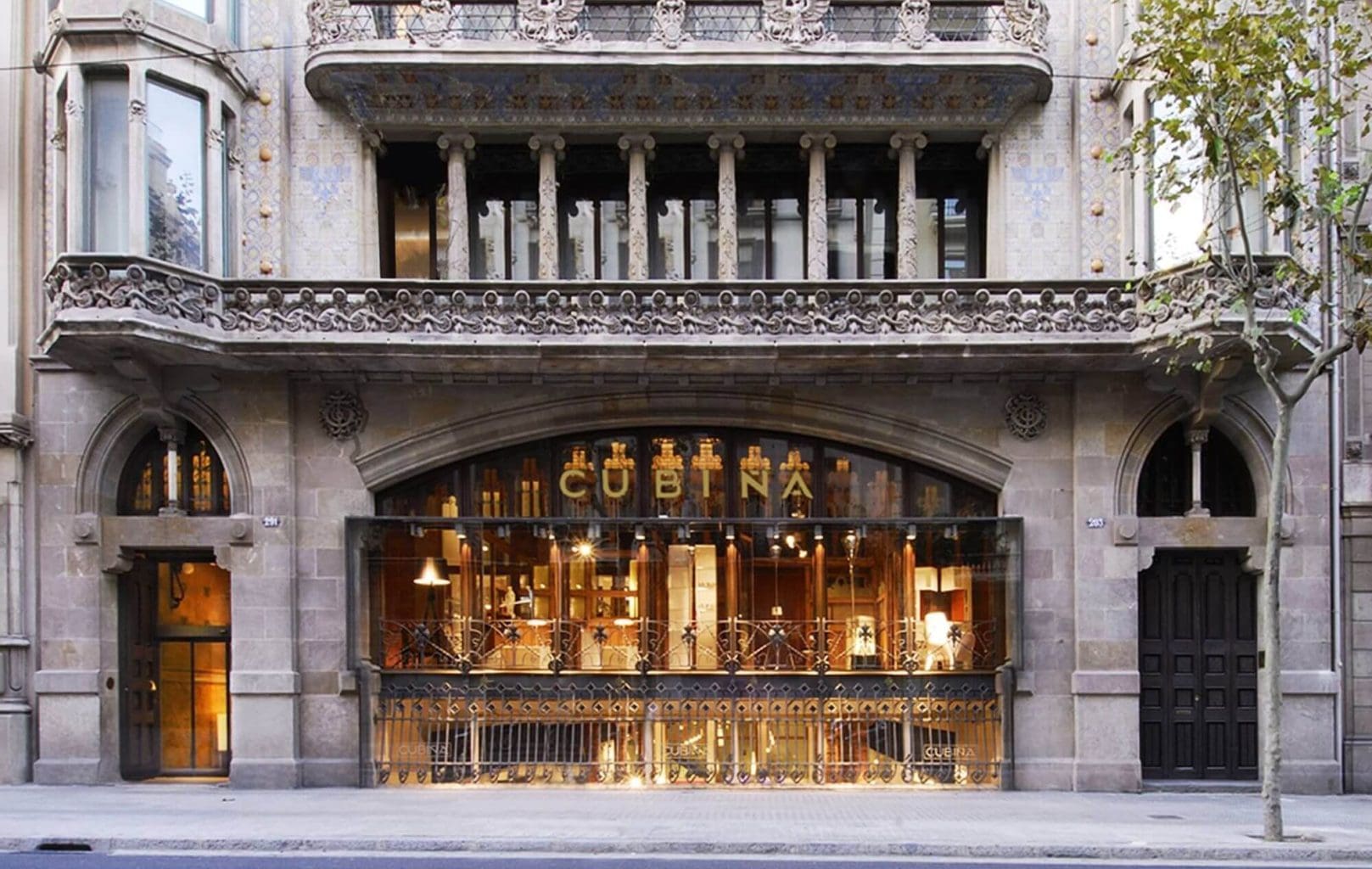 Street view of a historic stone building in Barcelona with large arched windows and a sign reading “CUBIÑA,” featuring decorative balconies, visible lighting inside, and reminiscent of the city's charming historic fortresses.