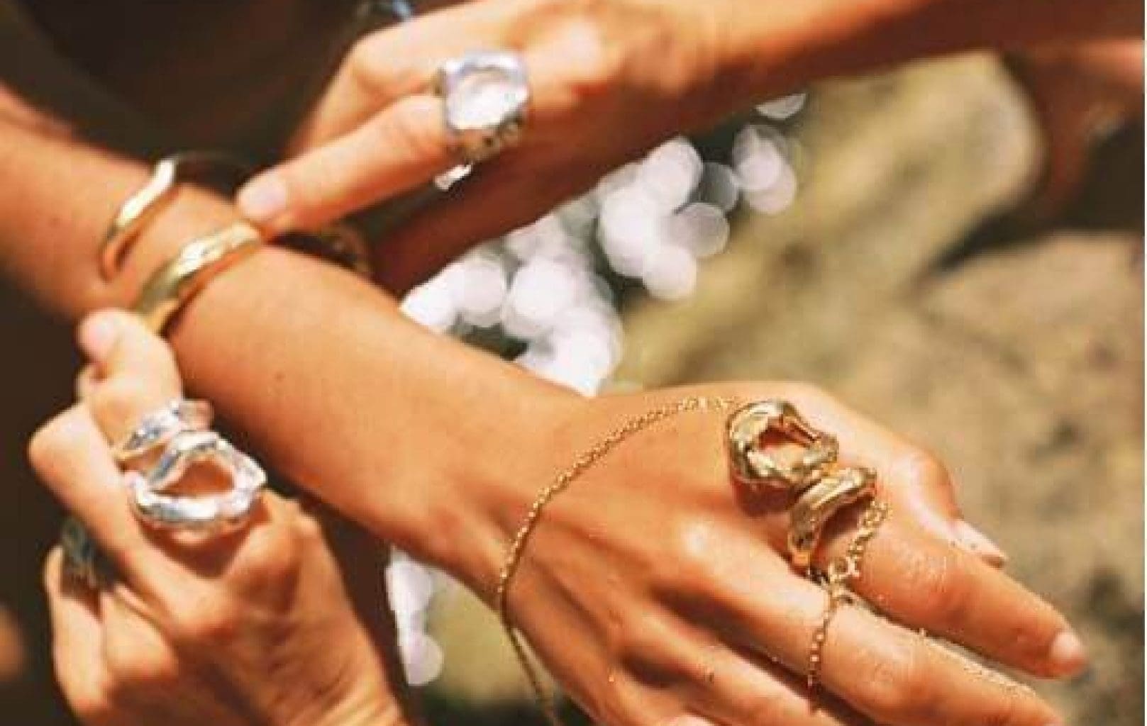 Varias manos exhiben gruesas pulseras y anillos de oro con piedras preciosas transparentes sobre un fondo exterior difuminado.