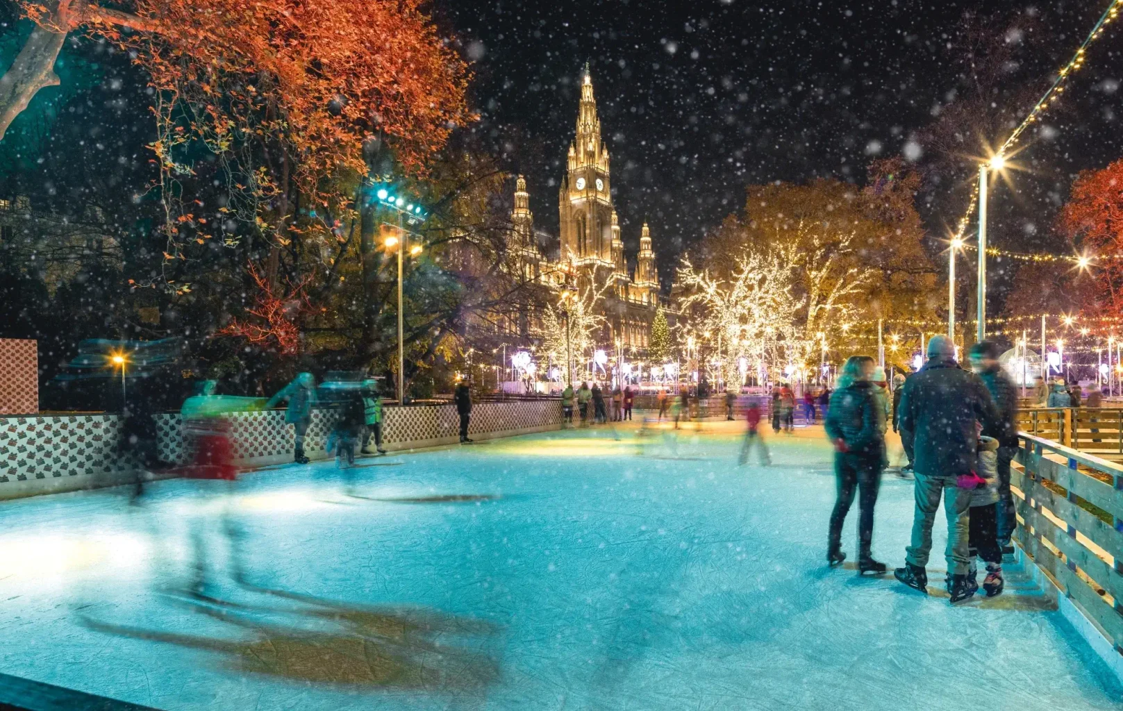 People ice skating at night in a festive outdoor rink at Vienna’s Christmas Markets, with trees decorated with lights and a large illuminated building in the background. Snow is falling gently.