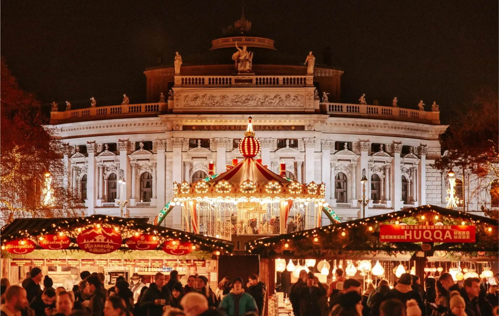A festive outdoor Christmas market at night with illuminated stalls, a carousel, and a grand historic building in the background crowded with people, evoking the magical charm of Vienna’s renowned Christmas Markets.