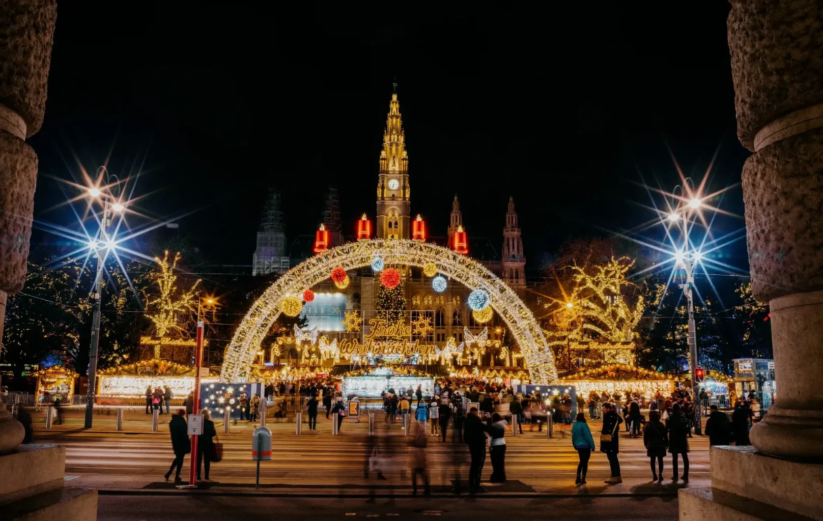 Crowds gather at Vienna’s brightly lit Christmas market, with a large illuminated arch, festive decorations, and a historic building in the background at night.