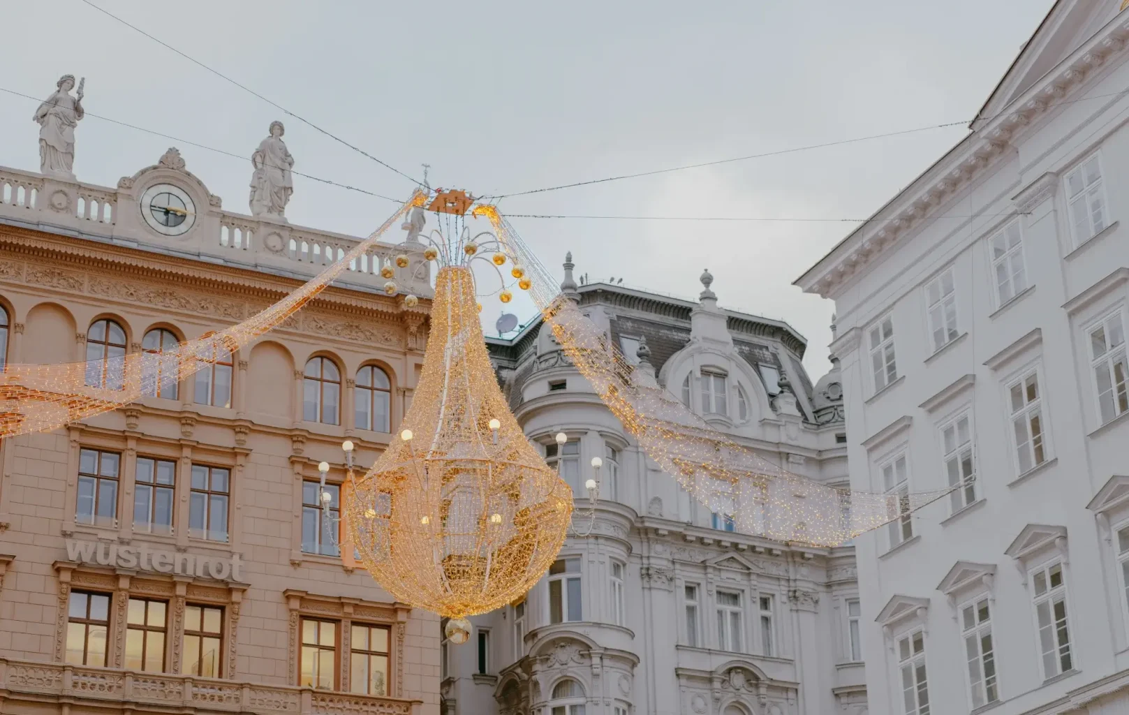 A large decorative chandelier-style light fixture hangs between ornate historic buildings in Vienna, with strings of lights and statues visible on the rooftops, capturing the festive spirit of Vienna Christmas Markets.