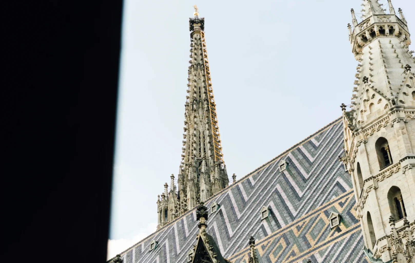 A section of St. Stephens Cathedral with a patterned, colorful tiled roof and pointed spire, partially framed by a dark foreground.