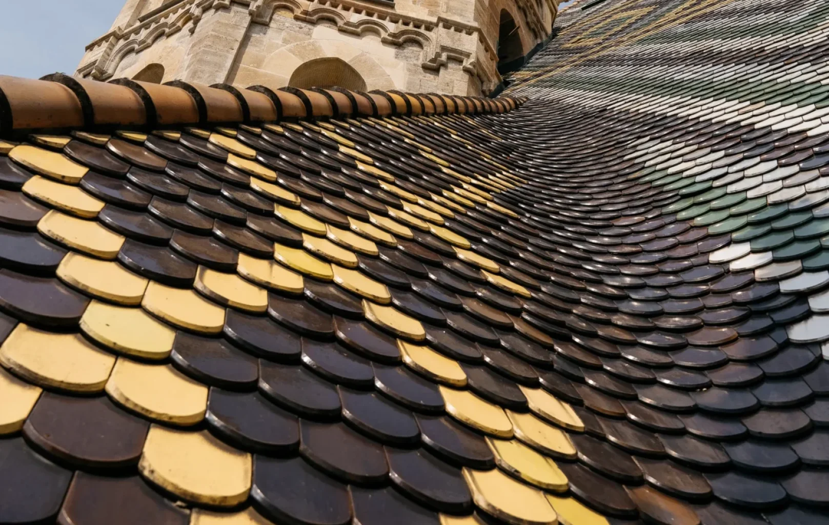 The stone church tower of St. Stephens Cathedral rises above a sloped roof covered in patterned, overlapping yellow and brown tiles under a partly cloudy Vienna sky.