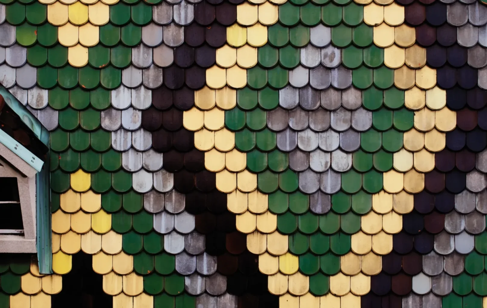 Close-up of the patterned roof at St. Stephens Cathedral in Vienna, showing diamond shapes in green, yellow, black, and gray tiles; part of a small dormer window is visible on the left.