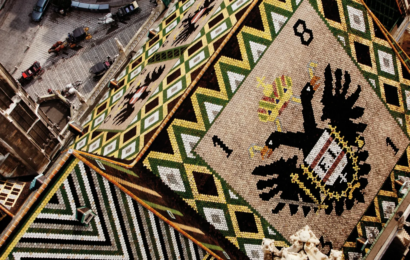 A close-up view of St. Stephens Cathedral’s colorful, tiled roof with geometric patterns and a black double-headed eagle emblem, overlooking a city square below.