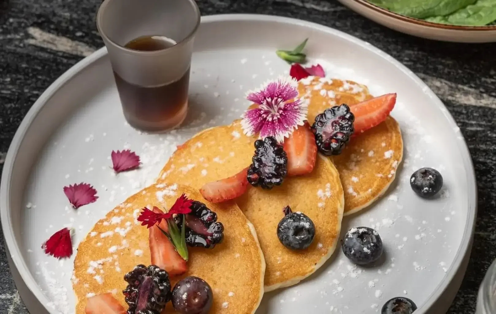 Tres tortitas cubiertas con fresas, moras, arándanos, flores comestibles y azúcar en polvo, servidas con un vasito de sirope en un plato blanco redondo.