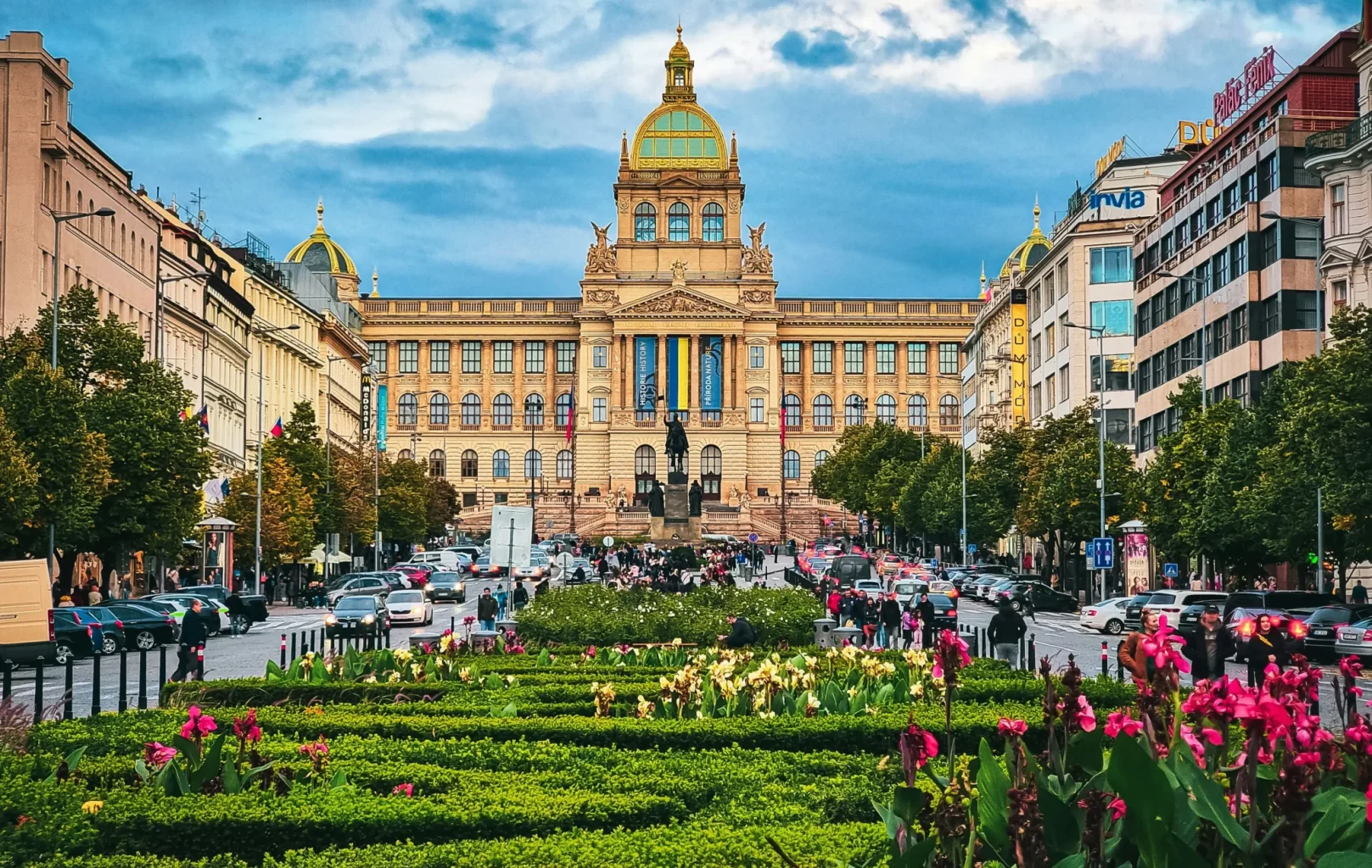 A large ornate building with a green dome stands at the end of Prague’s bustling Wenceslas Square, its boulevard lined with cars, trees, and flower beds under a cloudy sky.