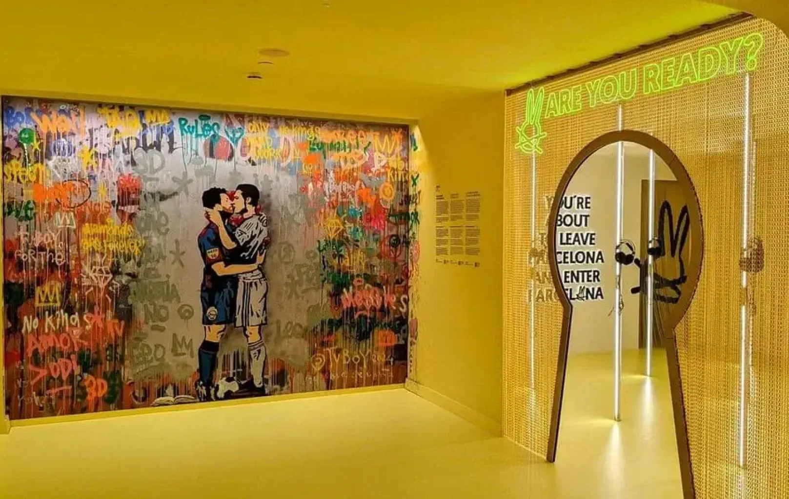 A yellow hallway at the White Rabbit Museum features a large graffiti-style mural of two soccer players kissing, with a neon "ARE YOU READY?" sign glowing above an arched doorway.