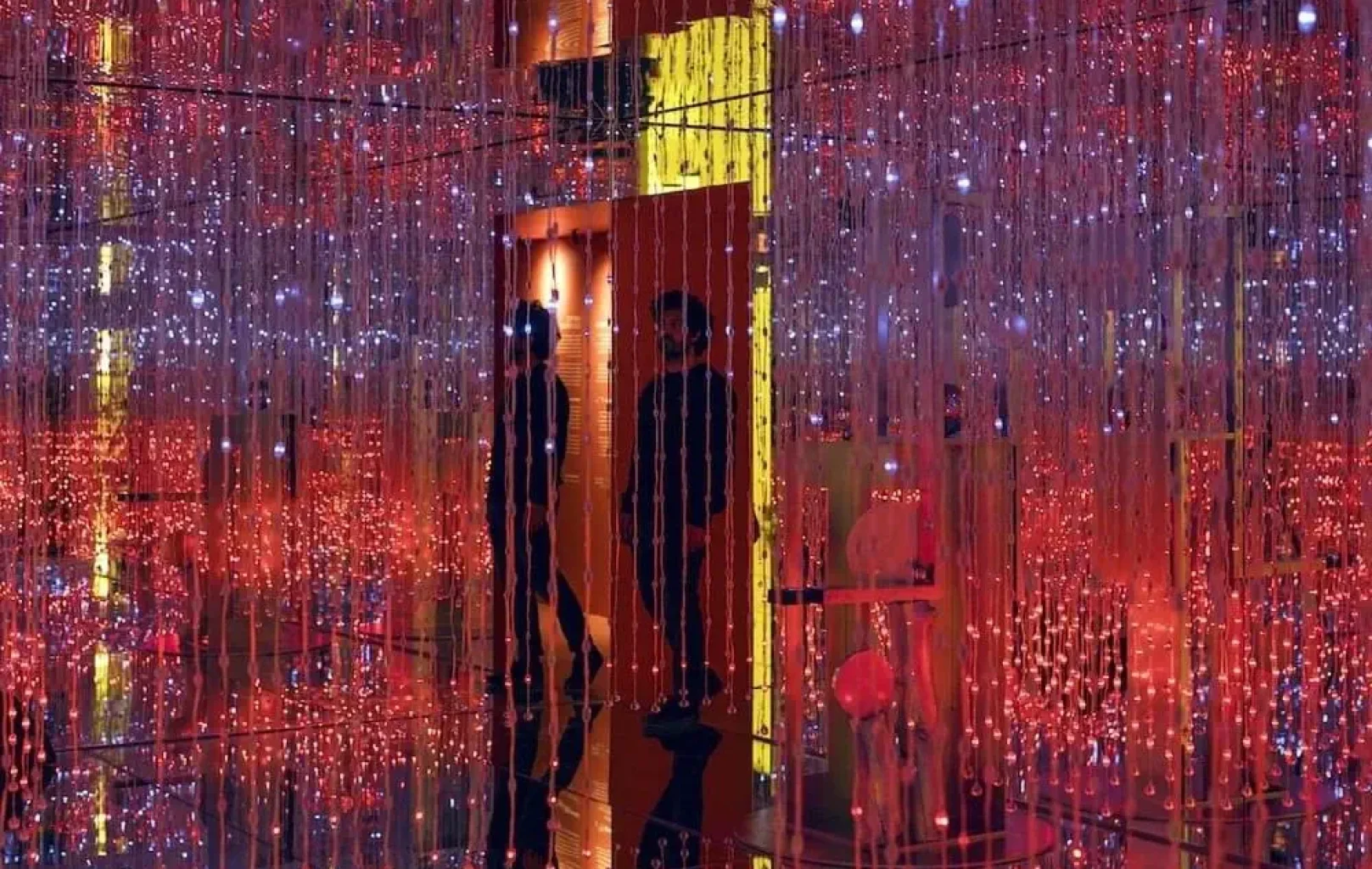 People stand among hanging strings of red and white LED lights in an immersive, mirrored art installation at the White Rabbit Museum.